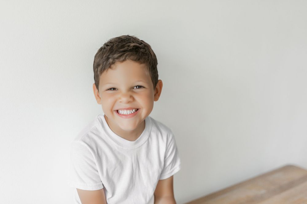 A young child in a white t-shirt smiles while posing for a portrait against a neutral background.