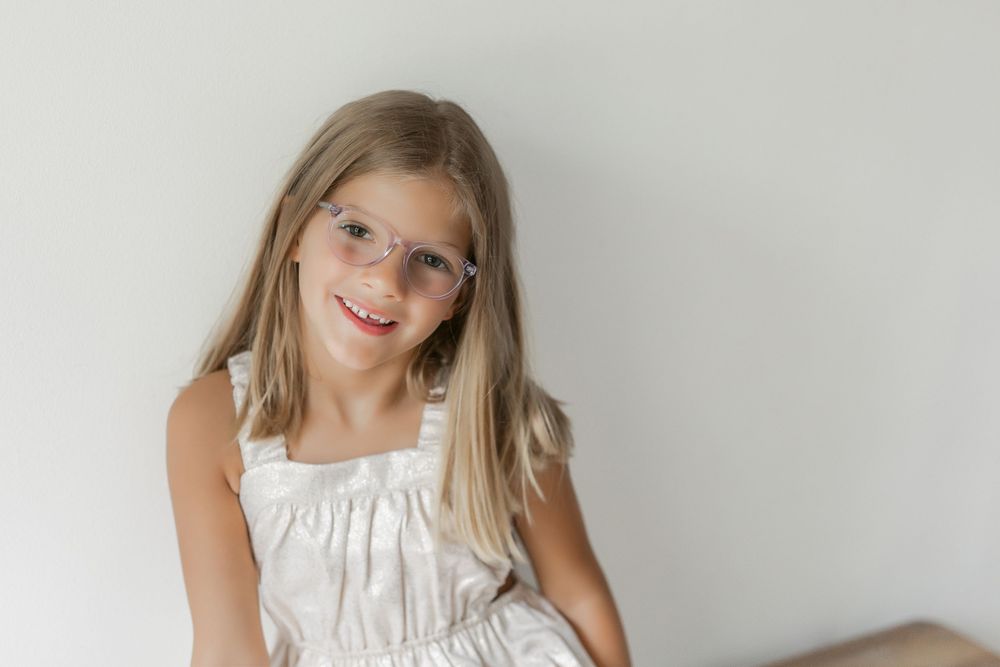 Young girl wearing a white ruffled dress poses against a plain wall with a bright smile.