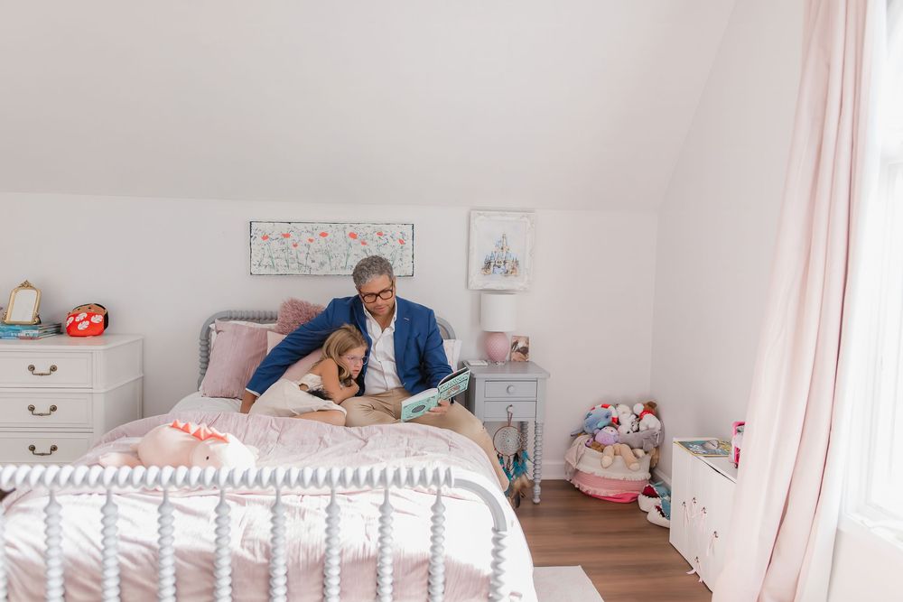 Two people share a sweet moment lying on a white bed in a bright, cozy bedroom with pink accents and decor.