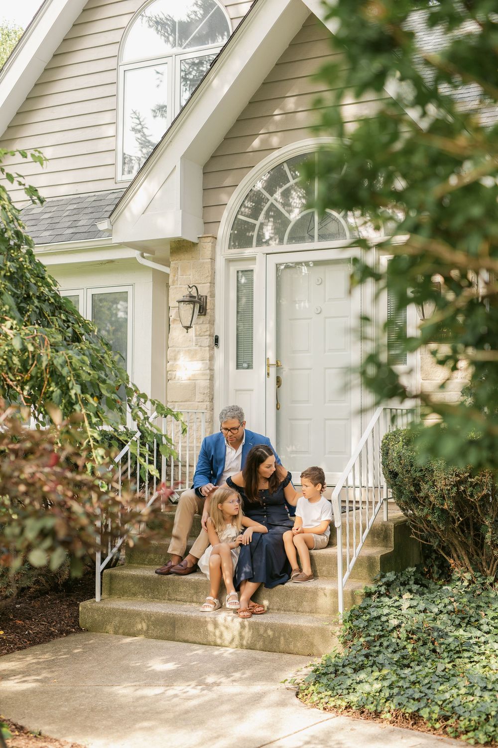 Family sitting together on the front steps of a white house with green bushes in summer.