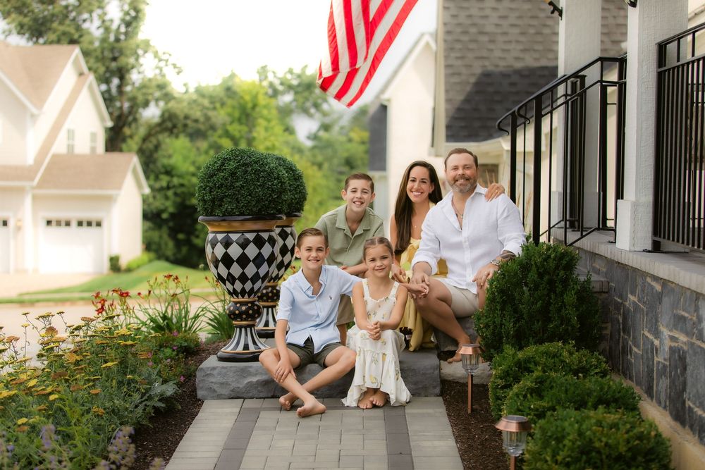 A family sits together on stone steps of their home porch with an American flag and landscaping visible.