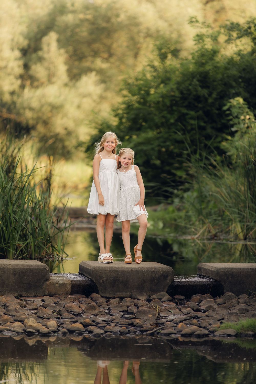 Two children in white dresses stand together on rocks by a peaceful pond reflecting the natural surroundings.