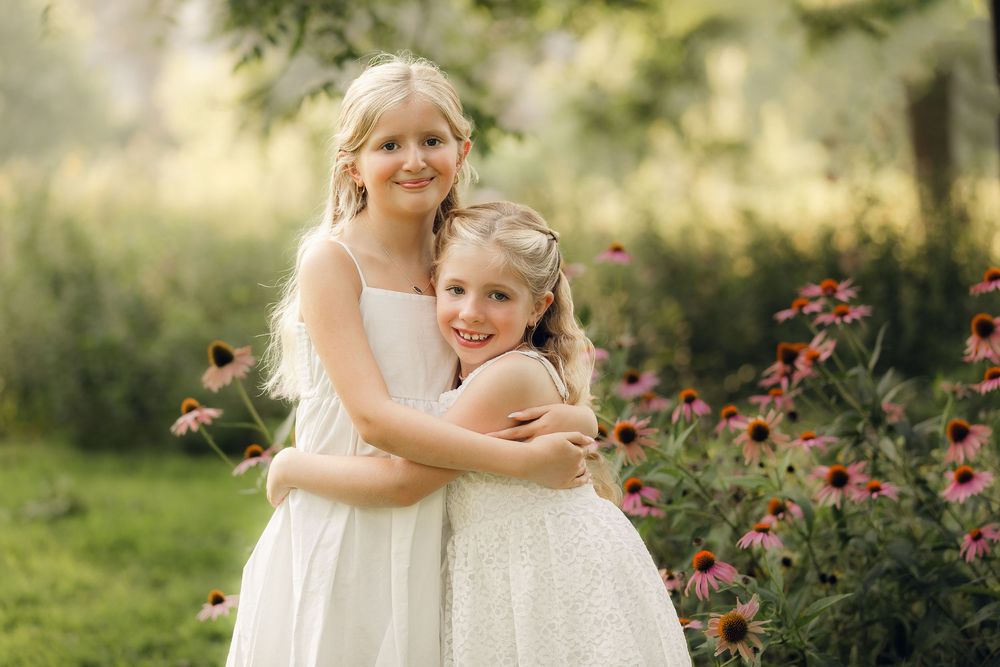 Two children in white dresses embrace while standing in a sunlit garden with pink flowers.