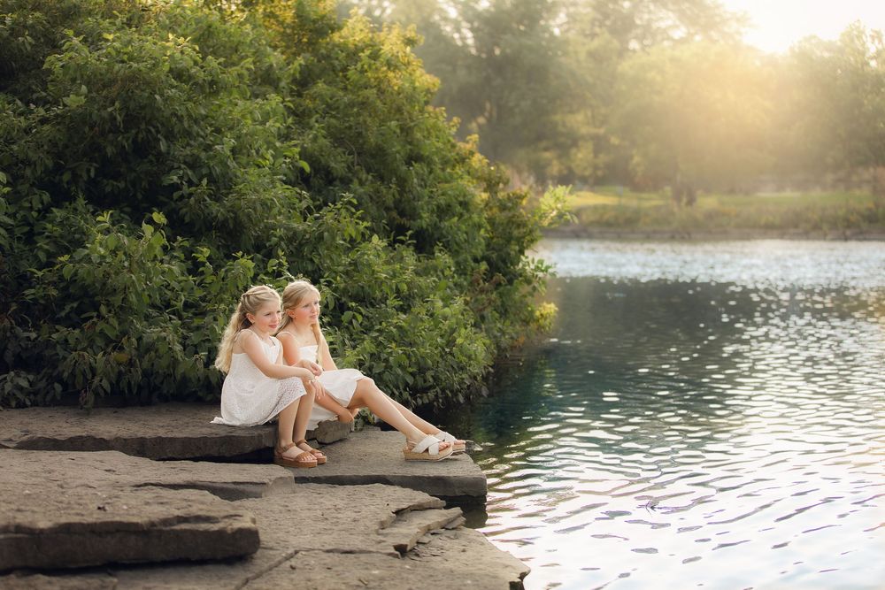Two people sit together on a rocky ledge by a tranquil lake at sunset while trees and golden light frame the scene.