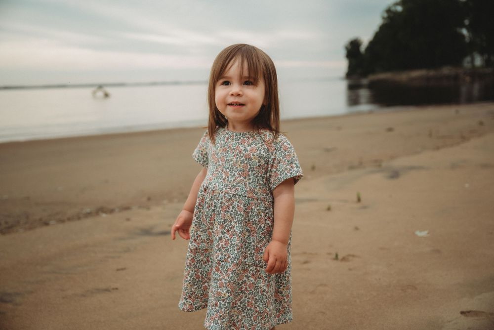 Young child in floral dress standing alone on beach at sunset.