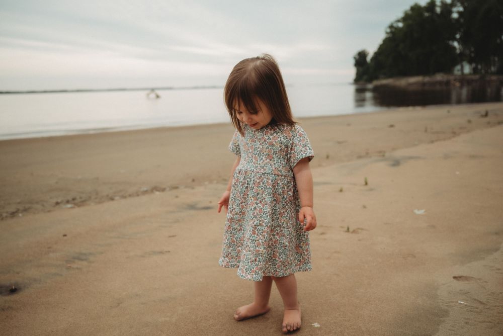Small child wearing patterned dress exploring sandy beach with cloudy skies.