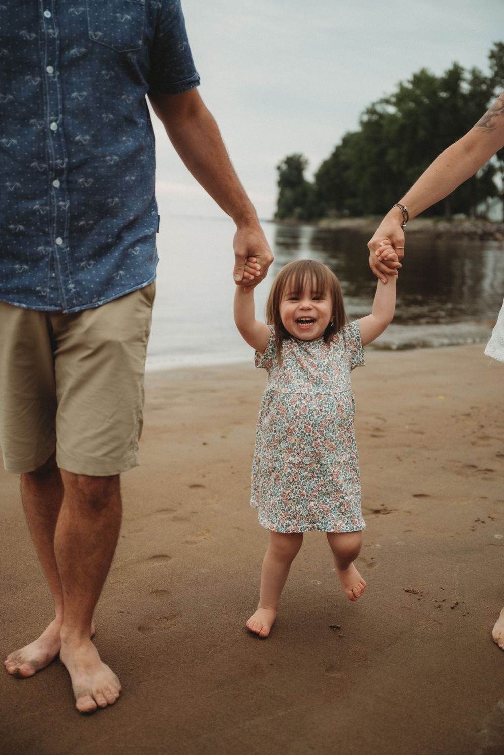 Child in floral dress joyfully swinging between adults on beach.