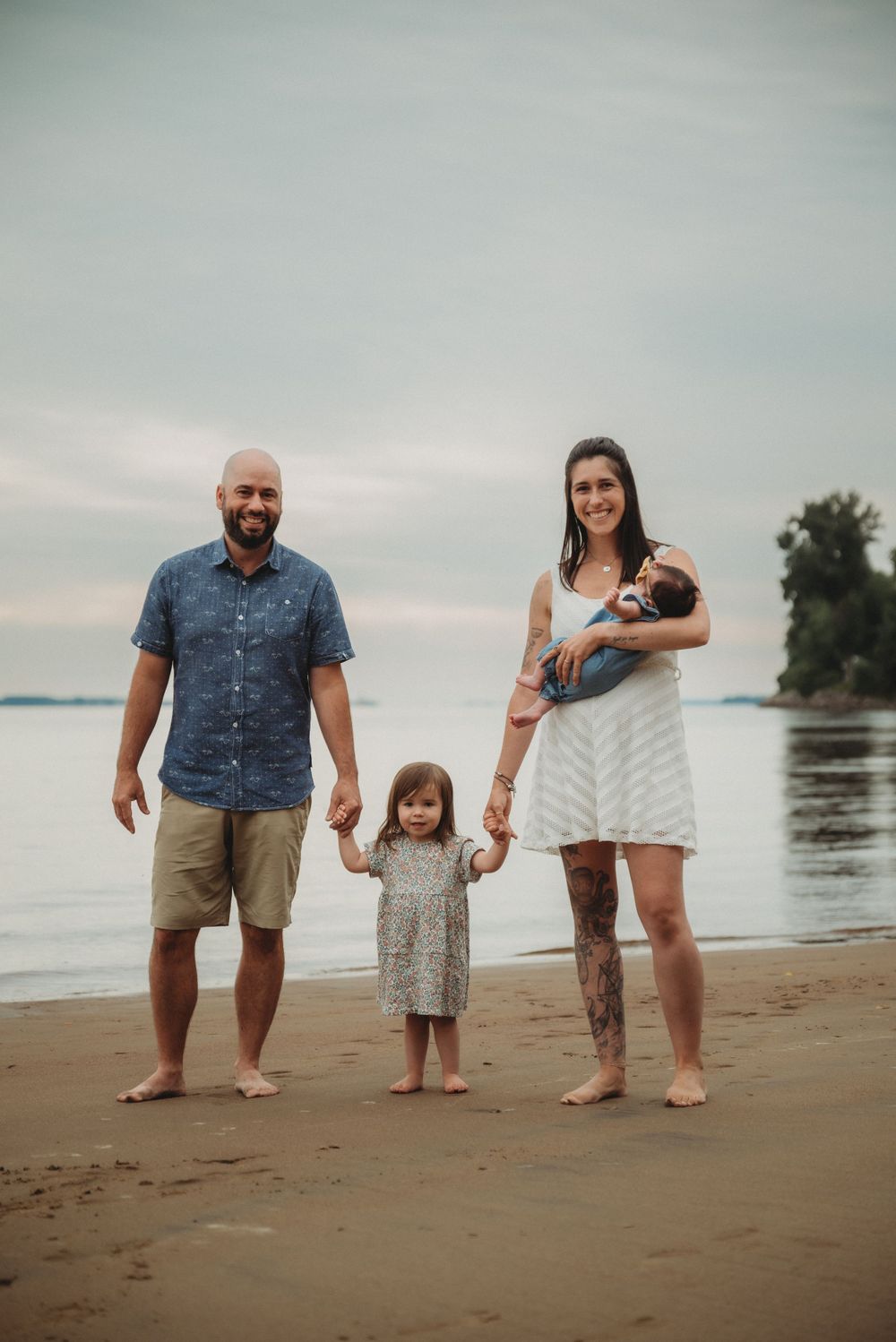 Family standing together on beach at sunset with water and trees in background.
