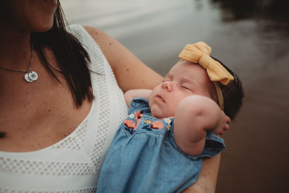 Baby wearing blue outfit with yellow bow sleeping peacefully against caregiver.