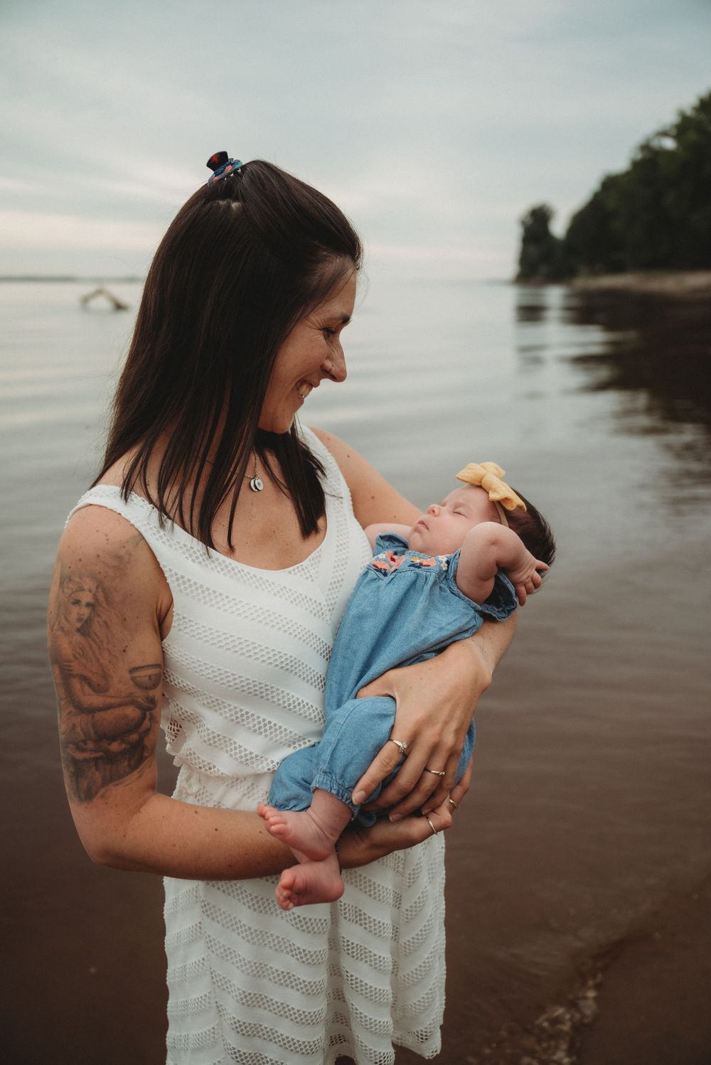 Person in white dress holding sleeping baby on beach at sunset.