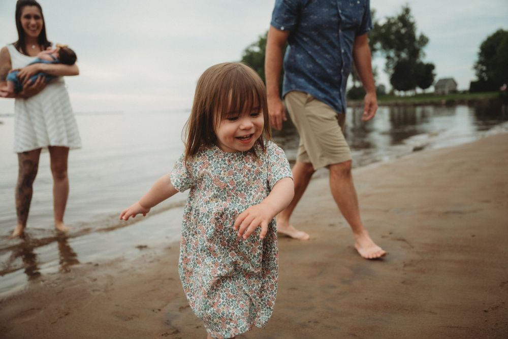 A small child runs joyfully along a beach at sunset while people watch from behind.