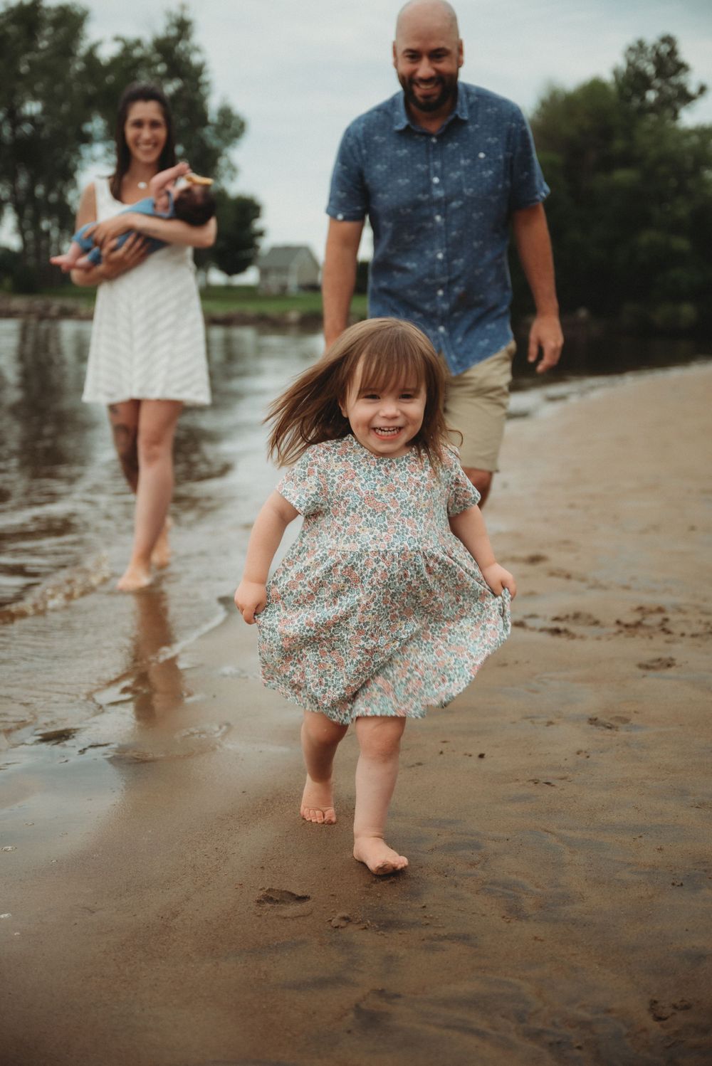 A toddler in a floral dress plays on a sandy beach while others stand nearby.