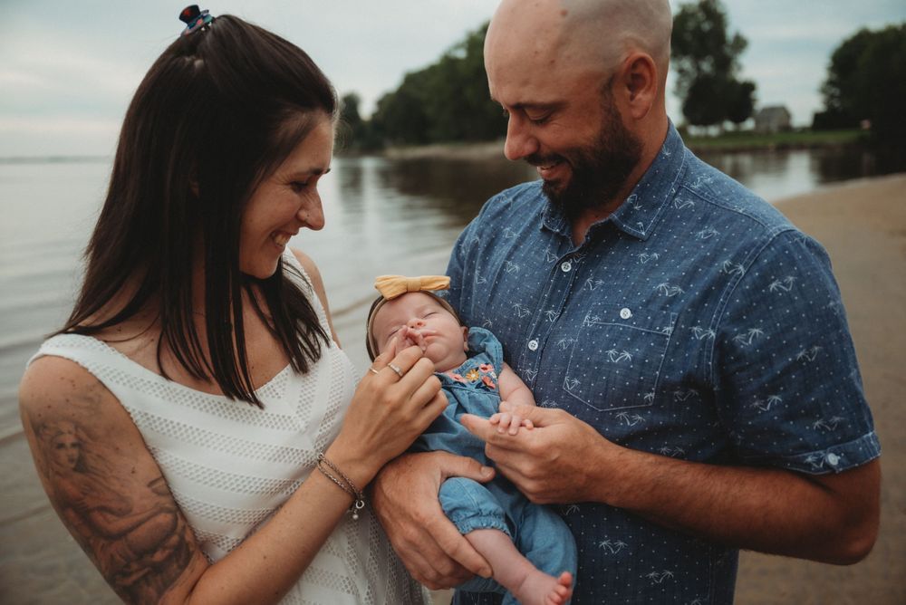 Parents holding their newborn baby by the waterfront on a cloudy day.