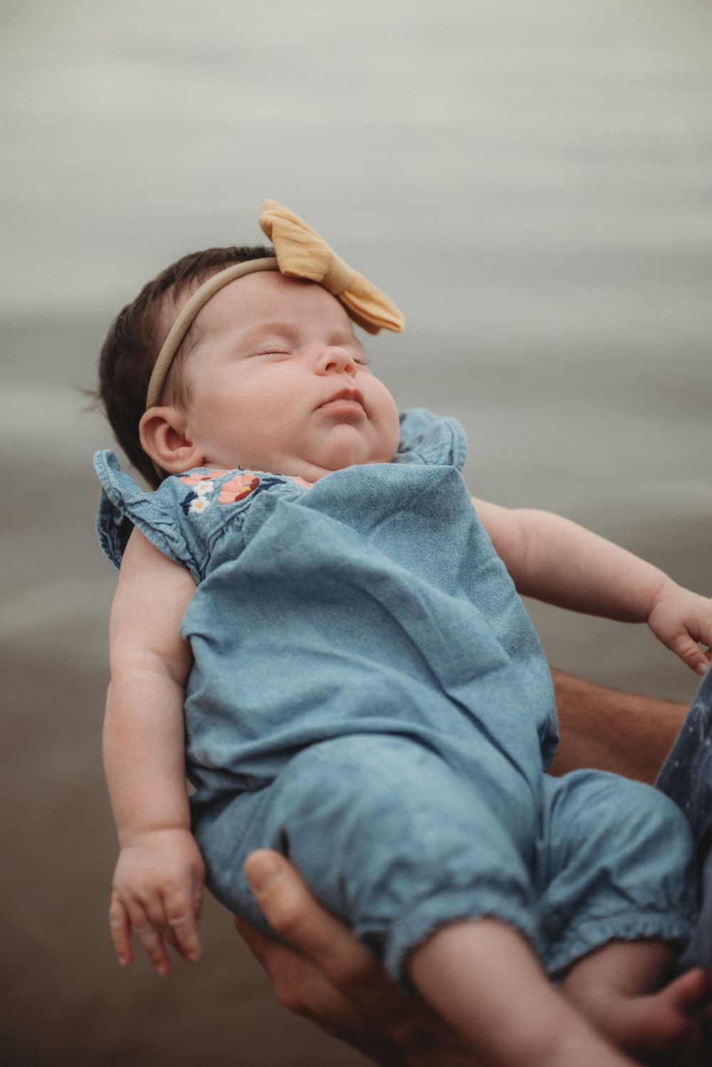 A sleeping baby wearing a blue romper and yellow bow headband rests peacefully.