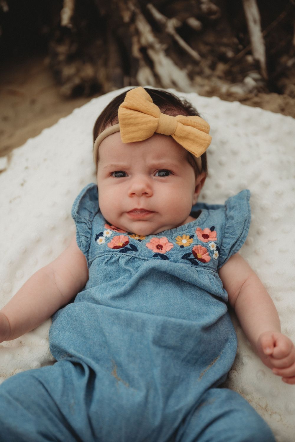 Small baby wearing blue floral romper and mustard bow headband laying on white blanket in natural setting.