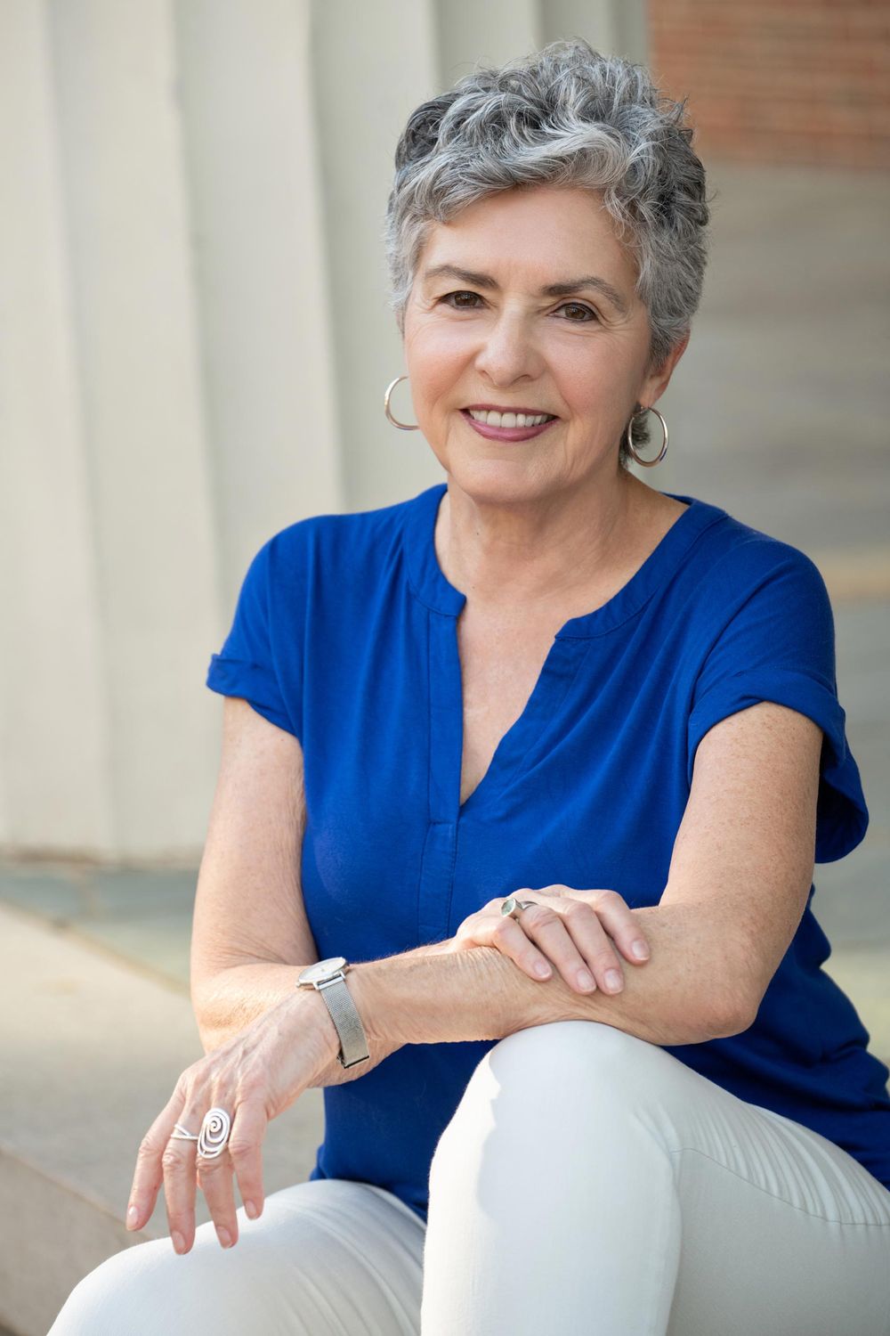 Professional headshot of a smiling woman in a blue top with silver hair sitting casually outside.