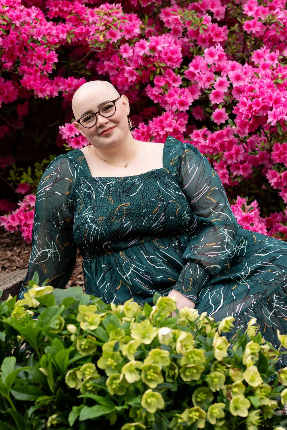 Person in a green patterned dress poses by vibrant pink azalea bushes and yellow-green shrubs in a garden setting.