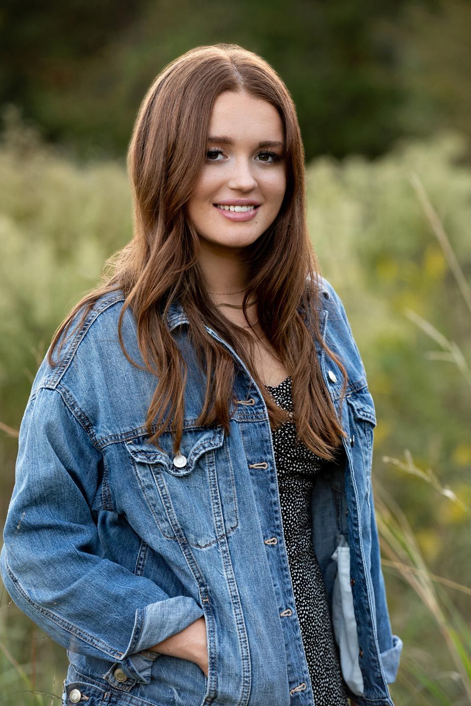 Person in distressed denim jacket poses outdoors with long brown hair against natural blurred background.