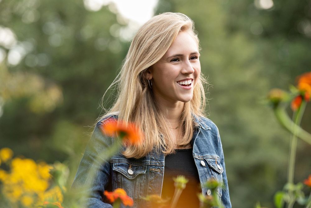 A person wearing a denim jacket smiles while standing in a garden with vibrant orange and yellow flowers.