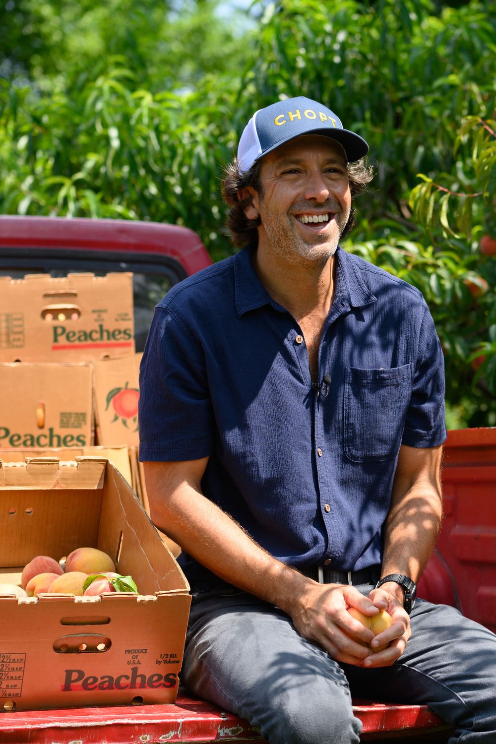 A person in navy shirt and baseball cap sits on a red truck bed with boxes of fresh peaches at a farm.
