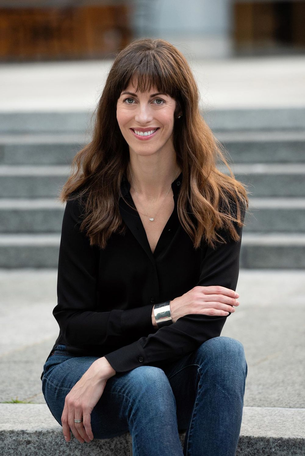 Professional woman in black blouse and jeans sitting casually on concrete steps with a warm smile.