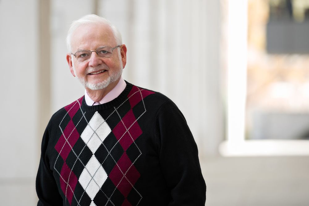 A person wearing a black and burgundy argyle sweater stands smiling in front of a light colored wall.