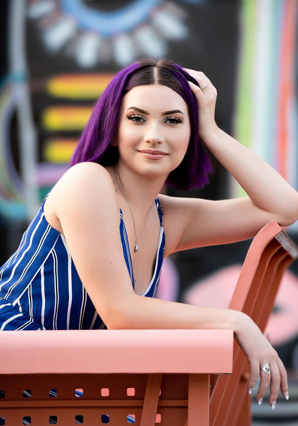 Woman in blue and white striped blazer poses on pink bench with purple hair highlights in outdoor setting.