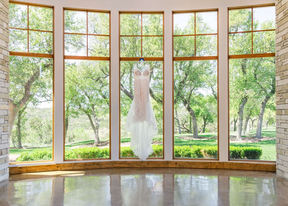 A white wedding dress hangs in front of large bay windows overlooking a green garden landscape.