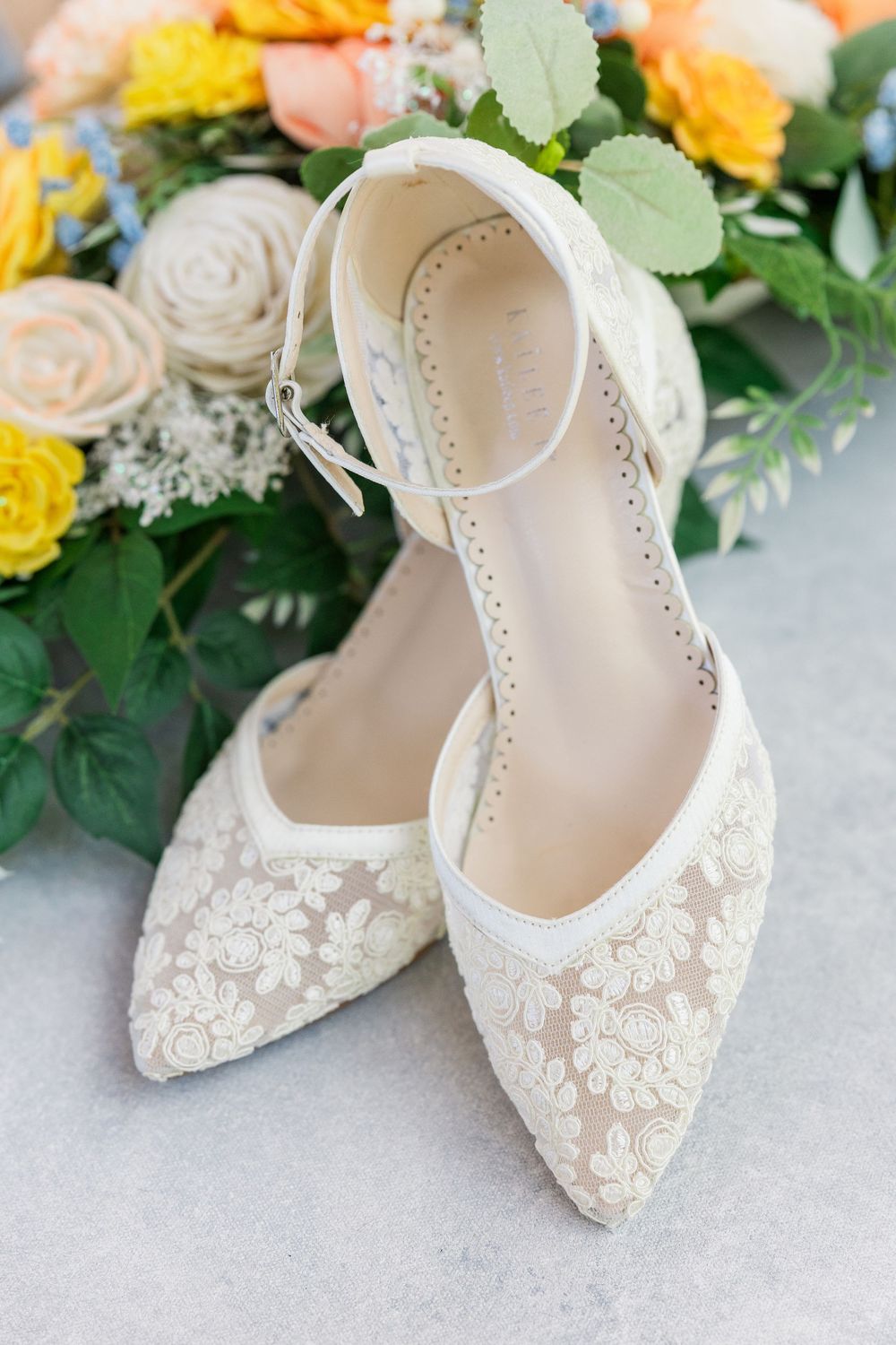 Elegant white lace bridal shoes with ankle straps displayed beside yellow and peach floral arrangements.