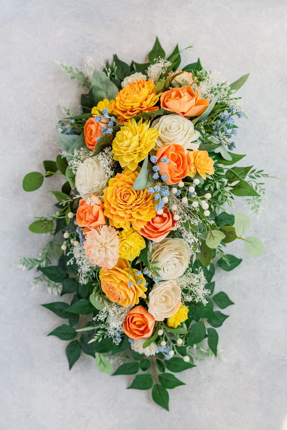 Elegant floral arrangement with orange, yellow and cream roses accented with baby's breath and green foliage on light grey background.