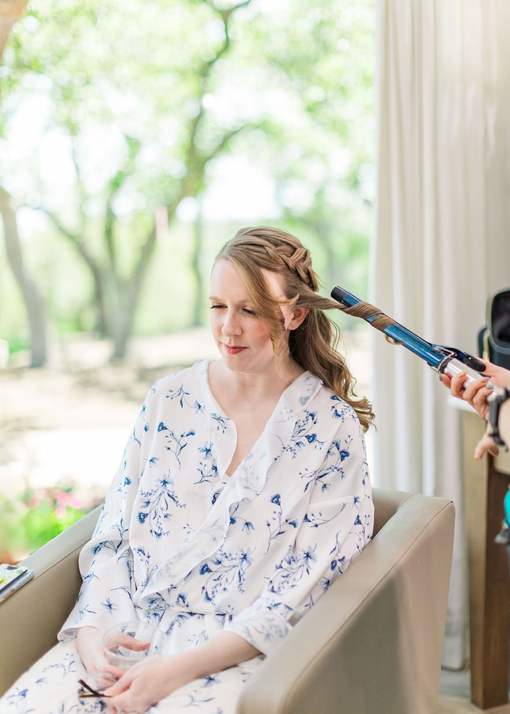 Getting ready photo of bride in floral robe having hair styled before wedding.