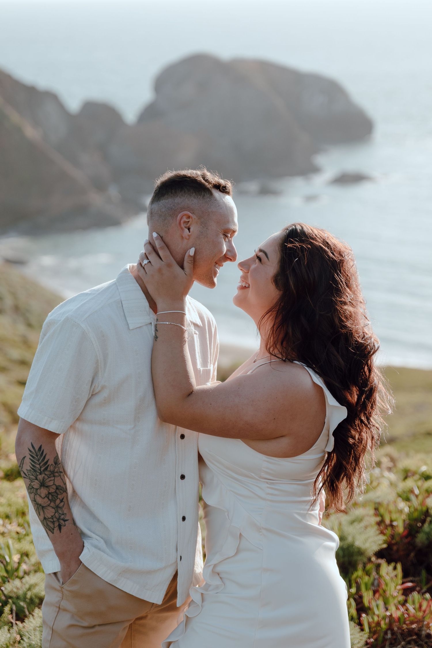 Romantic couple in white clothing share an intimate moment on a coastal cliffside with dramatic rock formations in background.
