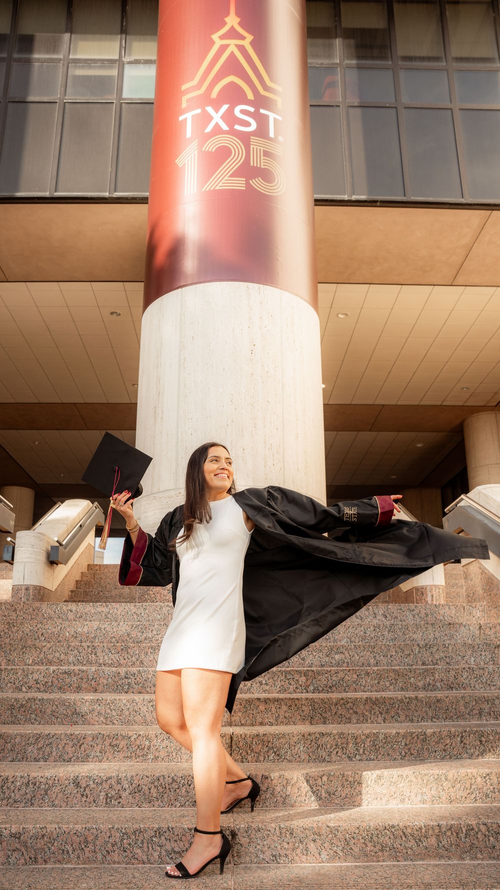 Graduate celebrates under TXST 125 sign on campus steps wearing cap and gown.