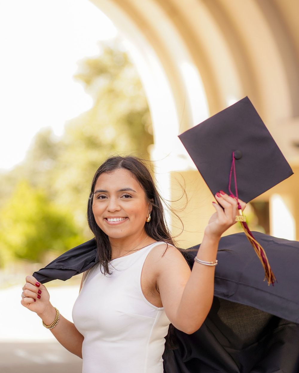 Graduate in white dress celebrates with cap and gown against architectural backdrop.