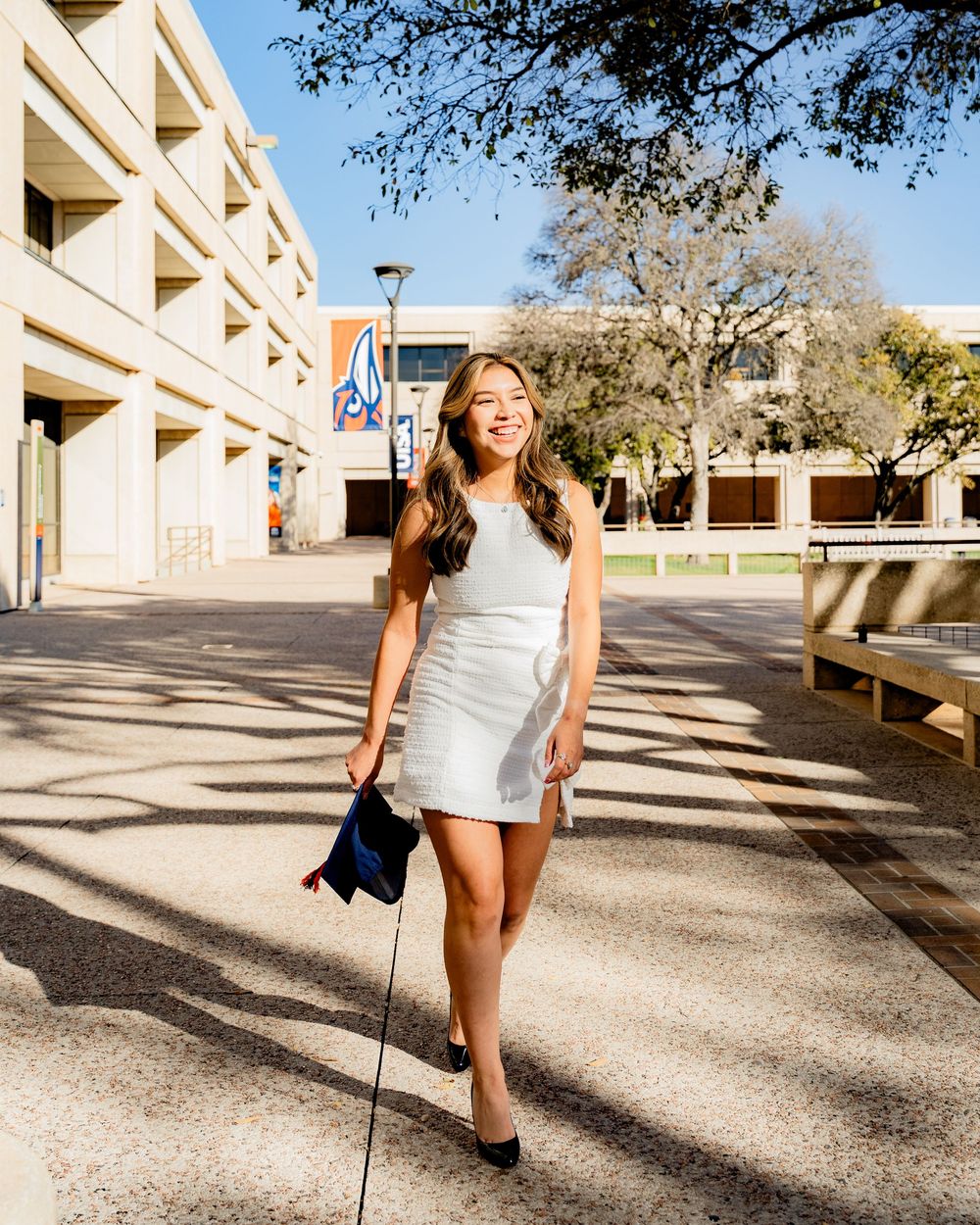 A person in a white dress walking across a sunny outdoor campus area.