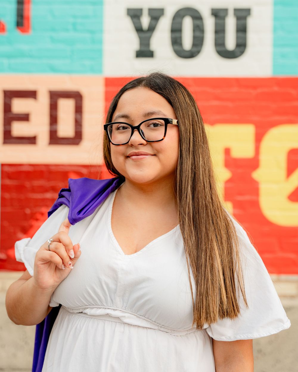 A student in white attire holds a purple backpack while standing in front of a colorful wall.