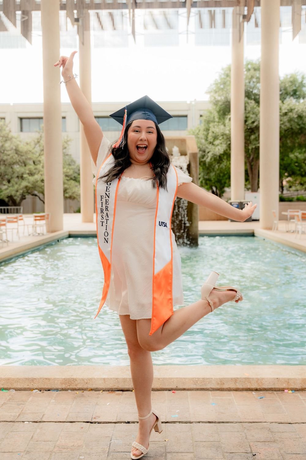 A graduate in cap and gown jumps joyfully in front of a fountain.