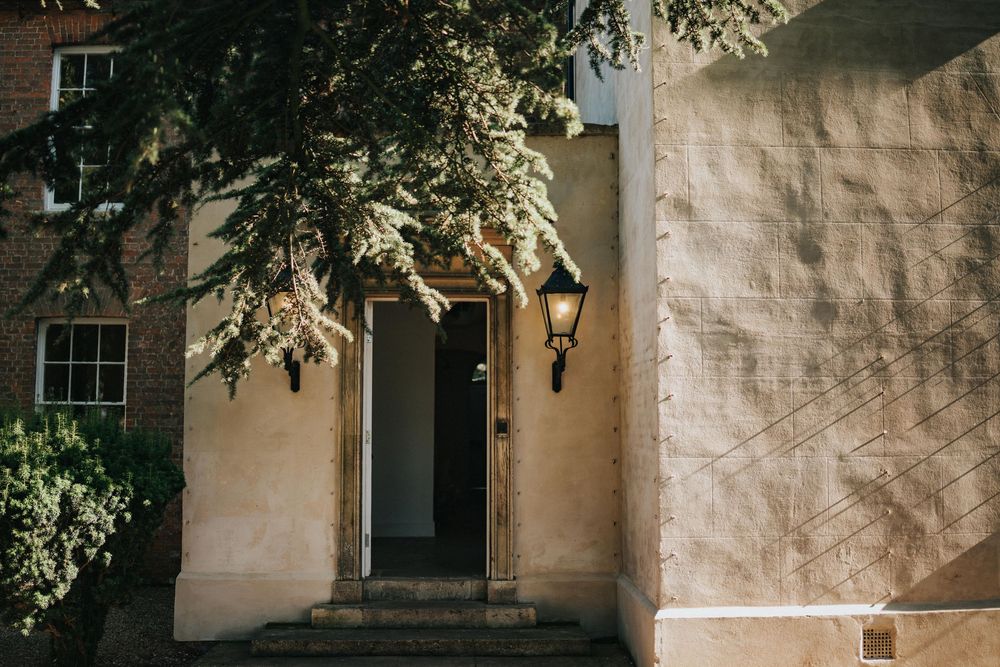 Vintage wall lantern decorates the entrance of a beige stucco house with a dark wooden door framed by green foliage at Aswarby Rectory.
