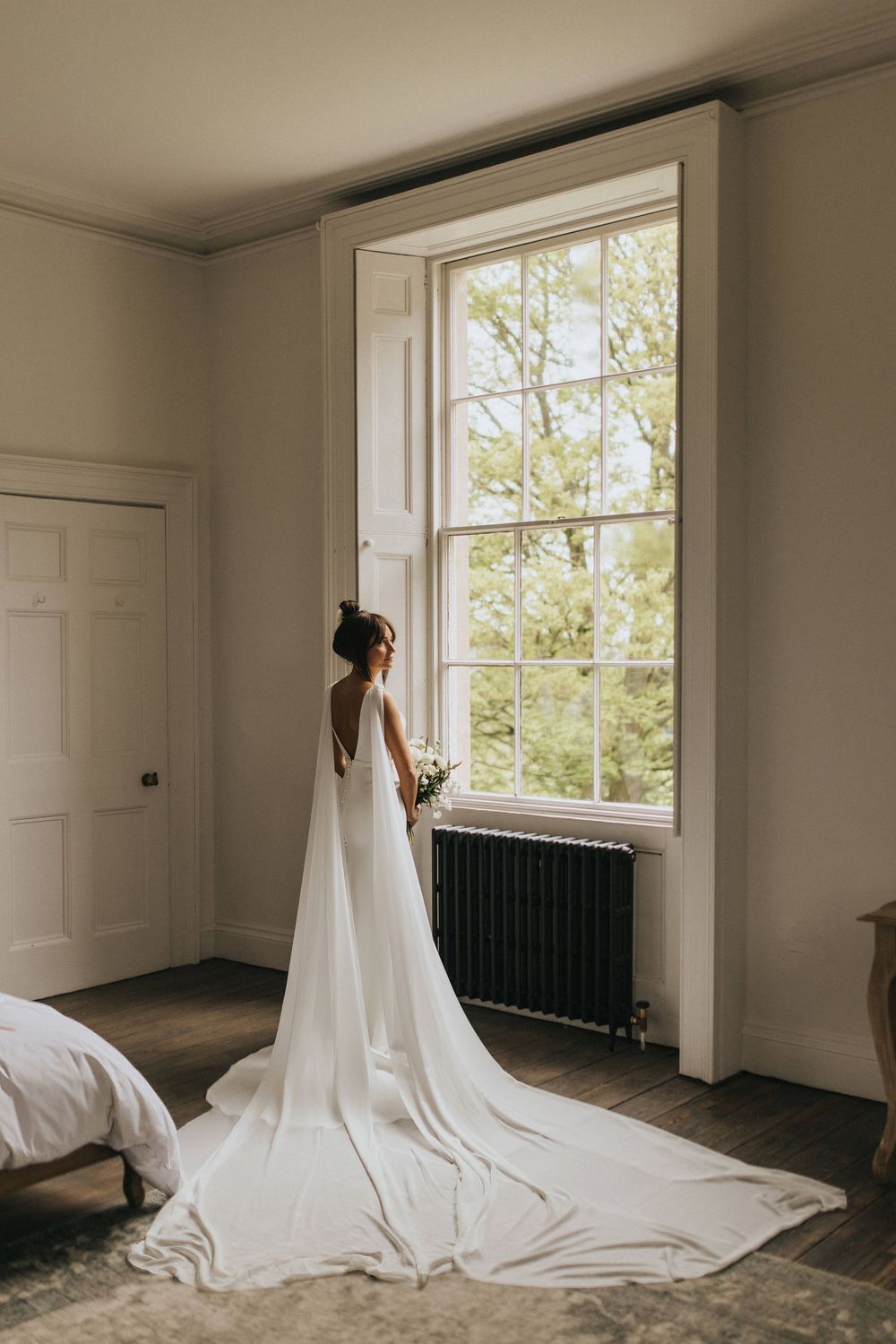 A bride in a flowing white gown with long train stands at a large window in an elegant minimalist room with sunlight at Aswarby Rectory.