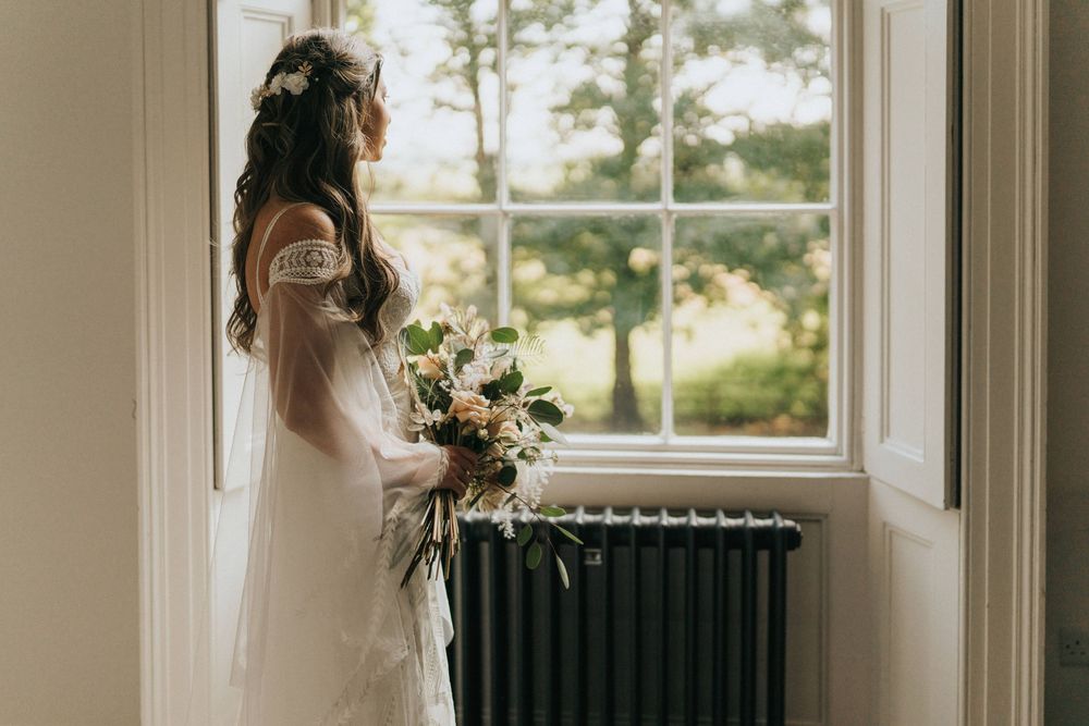 A bride in a white dress holds a delicate bouquet of flowers while standing by a window with a vintage radiator at Aswarby Rectory.