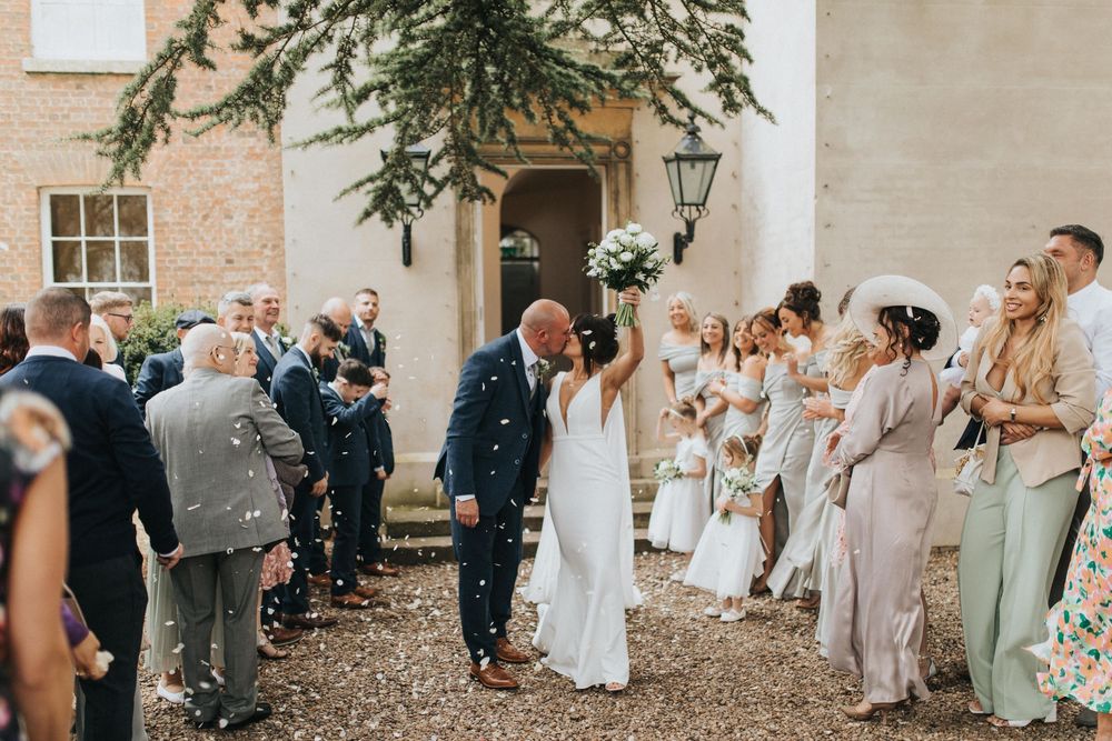 Wedding guests joyfully throw confetti at newlyweds outside a historic brick building at Aswarby Rectory.