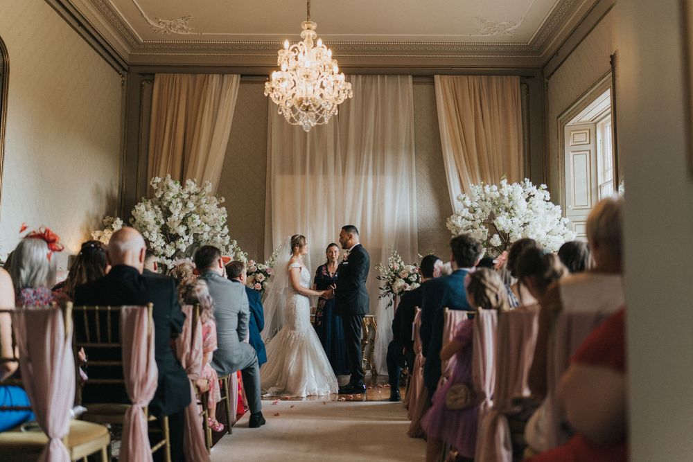 Elegant wedding ceremony takes place in a room with crystal chandelier and white floral arrangements at the altar at Rise Hall.
