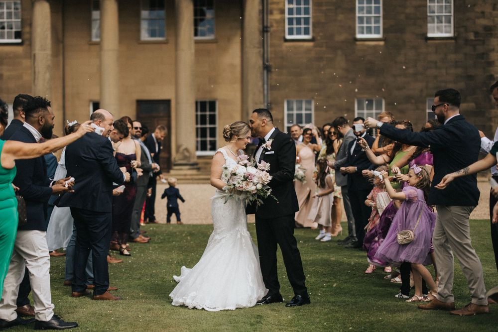 Newlyweds celebrate with guests throwing flower petals outside Rise Hall during wedding celebration.