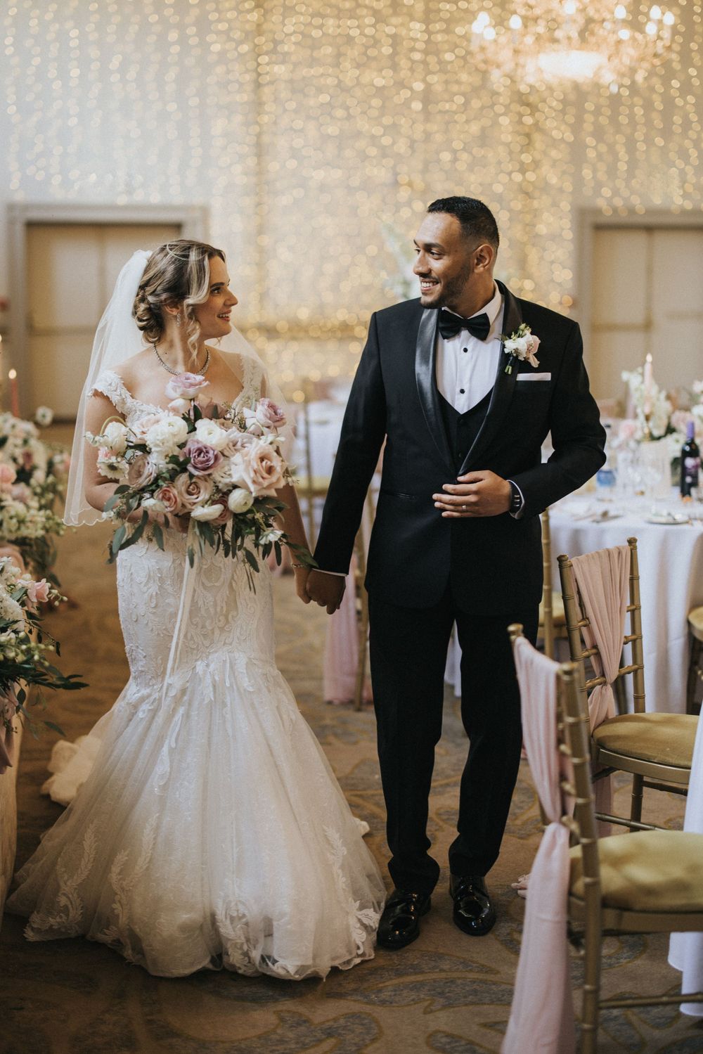 Elegant bride and groom walk together during their wedding reception in a beautifully lit ballroom with draped decor at Rise Hall.