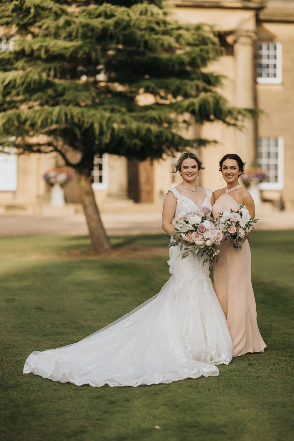 Two women in formal dresses stand on a manicured lawn holding floral bouquets at Rise Hall.