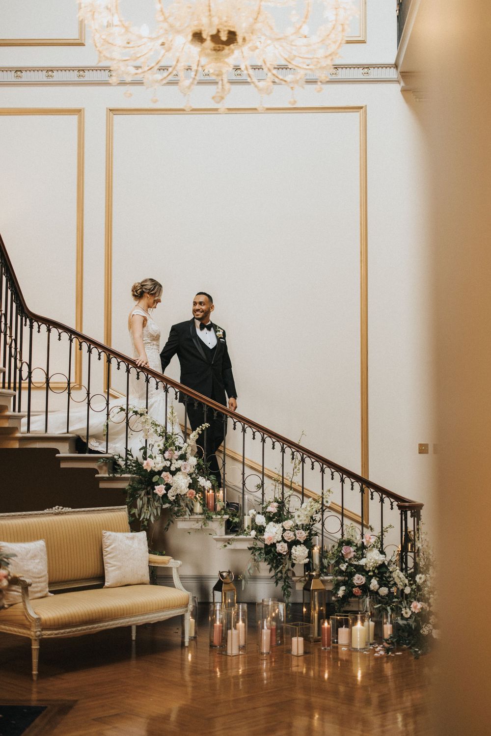 Bride and groom walk down a curved staircase adorned with white floral arrangements and romantic candlelight at Rise Hall.