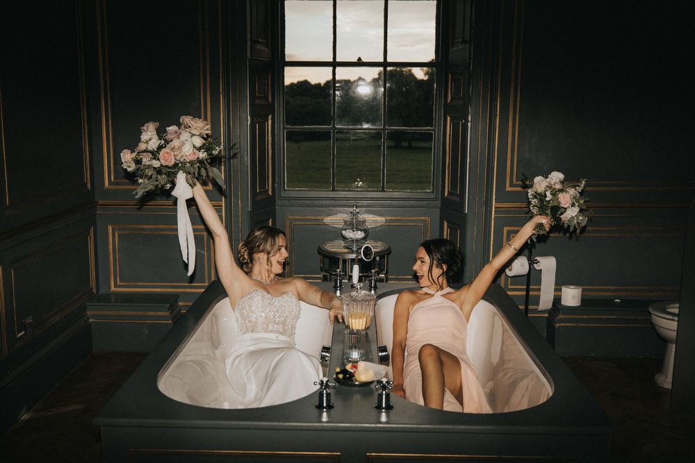 Bride and bridesmaid hold bouquets while relaxing in a luxurious bathroom with gold trim details at Rise Hall.