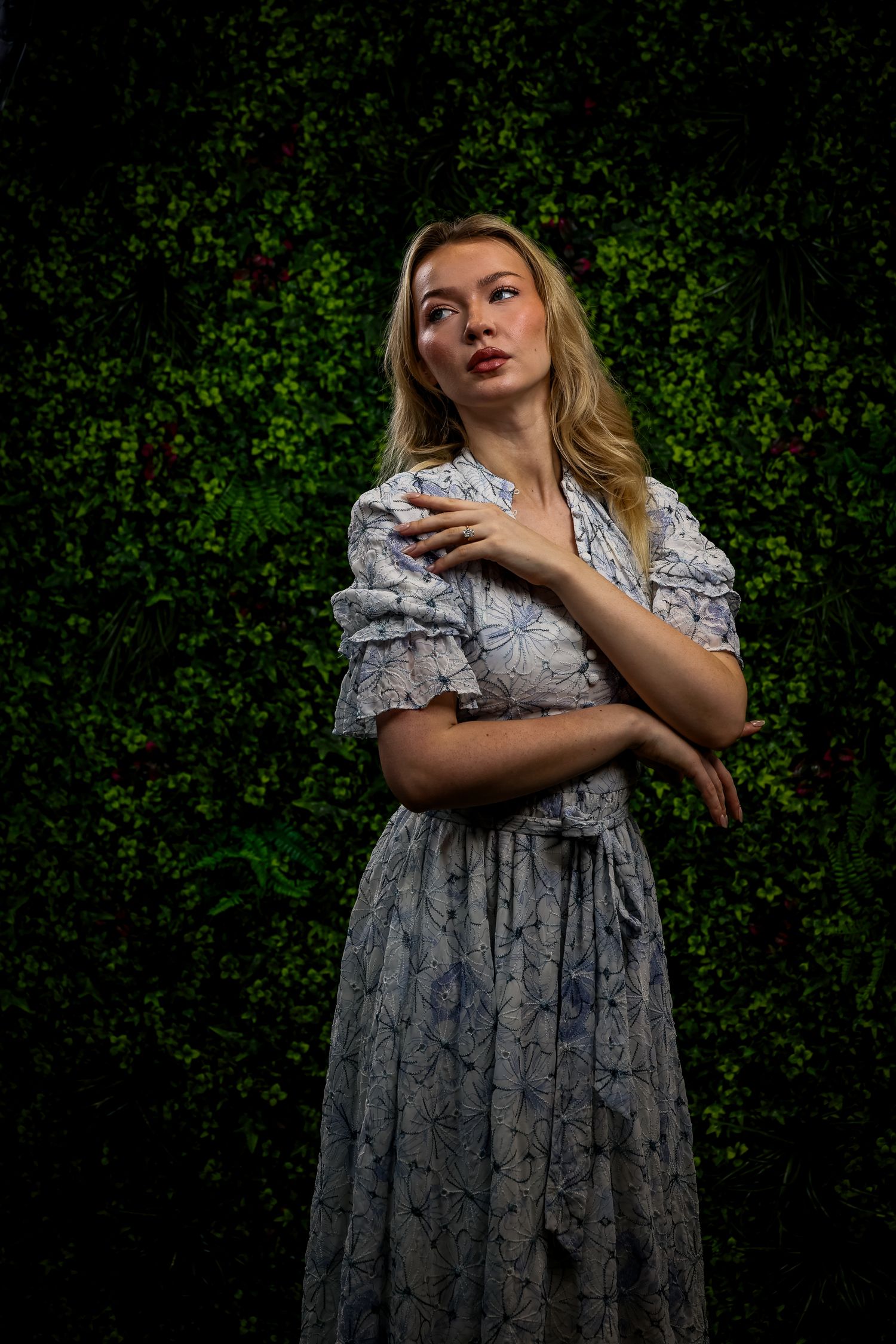 Woman in light floral dress poses artistically against dark green foliage background creating dramatic contrast.