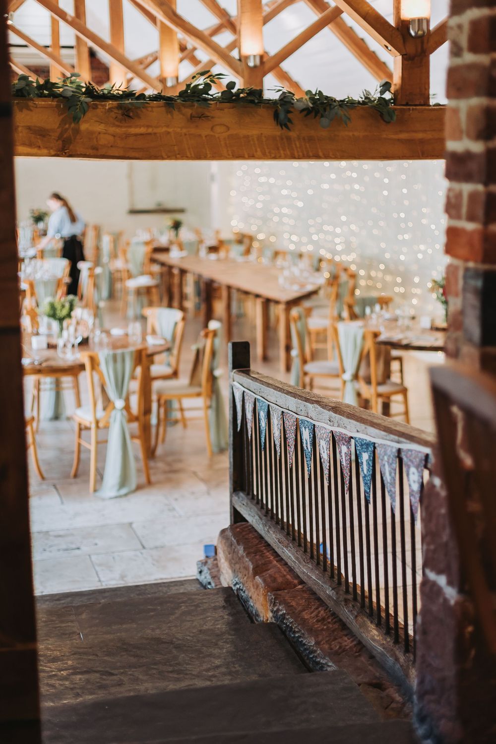 Wedding reception venue decorated with string lights and wooden beams features elegant dining tables and rustic decor.