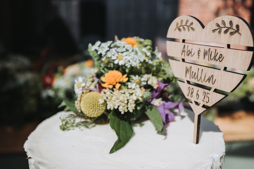 Rustic wooden heart-shaped wedding cake topper nestled among colorful wildflowers on a white frosted cake.